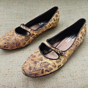 May include: A pair of women's flats featuring a leopard print pattern. The shoes have a rounded toe, a strap across the top, and brown trim. The flats are made of a textured fabric and are set on a neutral background.