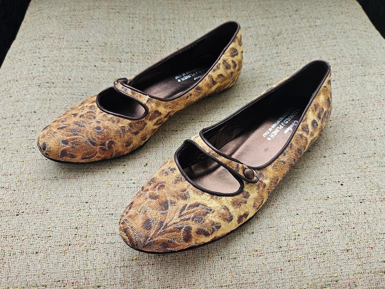 May include: A pair of women's flats featuring a leopard print pattern. The shoes have a rounded toe, a strap across the top, and brown trim. The flats are made of a textured fabric and are set on a neutral background.