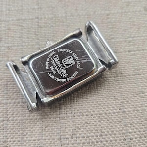 May include: A silver-toned watch back with the brand name "Olivia Rock" and the text "Stainless Steel Back" and "Quartz Movt". The watch back is rectangular and has two hinged clasps. The watch back is made in China.