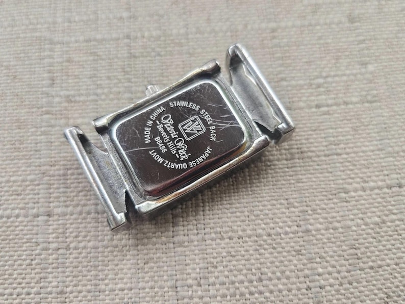 May include: A silver-toned watch back with the brand name "Olivia Rock" and the text "Stainless Steel Back" and "Quartz Movt". The watch back is rectangular and has two hinged clasps. The watch back is made in China.