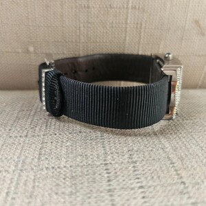 May include: A silver wristwatch with a black ribbed band and a dark brown leather backing. The watch face is rectangular and has a diamond-like embellishment. The watch is resting on a neutral-colored surface.