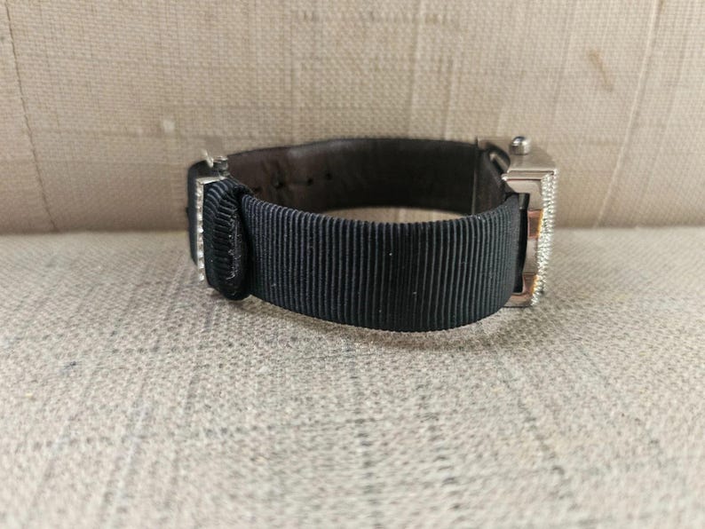 May include: A silver wristwatch with a black ribbed band and a dark brown leather backing. The watch face is rectangular and has a diamond-like embellishment. The watch is resting on a neutral-colored surface.