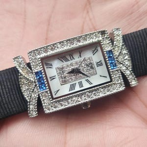 May include: A rectangular wristwatch with a silver-colored bezel encrusted with clear crystals and blue accents. The watch face is white with black Roman numerals. The watch is attached to a black fabric band.