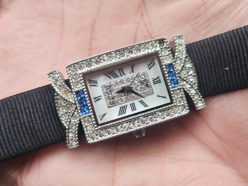 May include: A rectangular wristwatch with a silver-colored bezel encrusted with clear crystals and blue accents. The watch face is white with black Roman numerals. The watch is attached to a black fabric band.