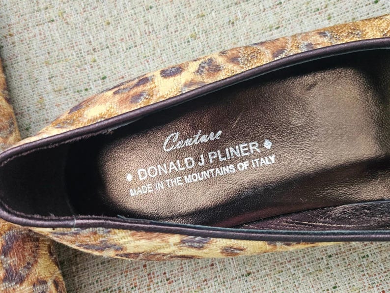 May include: Close-up of a shoe interior with a leopard print exterior. The shoe's insole is brown with the text "Couture DONALD J PLINER MADE IN THE MOUNTAINS OF ITALY". The shoe has a dark brown trim.