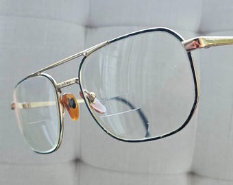 May include: Gold-framed spectacles with a double bridge and clear lenses. The frames have a classic aviator style with a rectangular shape. The nose pads are a light brown colour. The glasses are set against a neutral background.