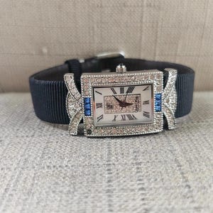 May include: A rectangular silver-toned watch with a white face and Roman numerals. The watch is adorned with clear and blue crystals and has a dark blue fabric band. The watch band is approximately 0.75 inches wide.