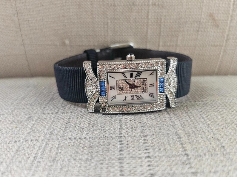May include: A rectangular silver-toned watch with a white face and Roman numerals. The watch is adorned with clear and blue crystals and has a dark blue fabric band. The watch band is approximately 0.75 inches wide.