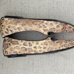 May include: A pair of flat shoes featuring a leopard print design. The shoes have a rounded toe and black trim. The color palette includes tan, brown, and black. The shoes are made from a textured material.
