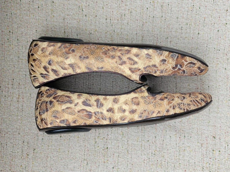 May include: A pair of flat shoes featuring a leopard print design. The shoes have a rounded toe and black trim. The color palette includes tan, brown, and black. The shoes are made from a textured material.