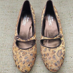 May include: A pair of leopard print flat shoes with pointed toes. The shoes have brown leather trim and a strap with a button fastening. The brand name "DONALD J PLINER" is visible. The shoes are made in Italy.