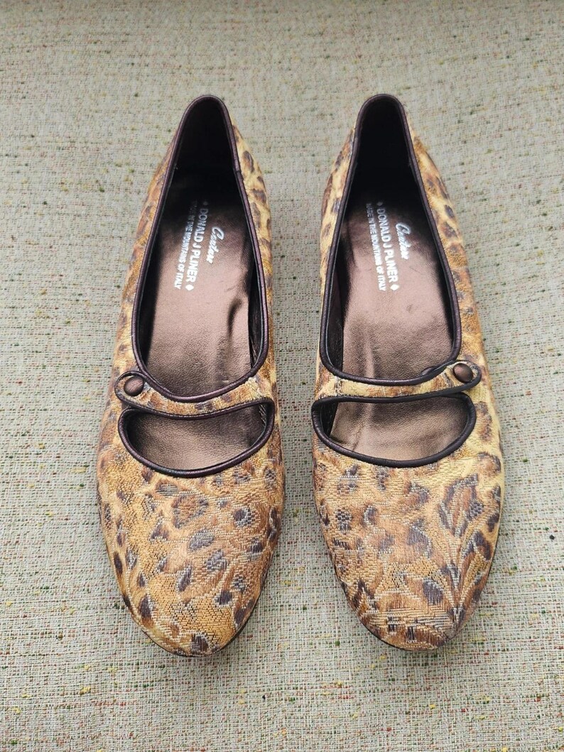 May include: A pair of leopard print flat shoes with pointed toes. The shoes have brown leather trim and a strap with a button fastening. The brand name "DONALD J PLINER" is visible. The shoes are made in Italy.