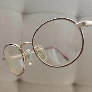 Może przedstawiać: Zbliżenie okularów w złotej oprawie z okrągłymi soczewkami. Okulary mają delikatny, minimalistyczny design, charakteryzujący się cienką, złotą oprawą i przezroczystymi soczewkami. Noski są w kolorze jasnozielonym. Ogólna estetyka jest prosta i elegancka.
