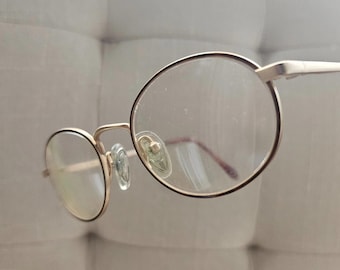 Puede incluir: Primer plano de gafas con montura dorada y lentes redondas. Las gafas tienen un diseño delicado y minimalista, con una fina montura dorada y lentes transparentes. Las almohadillas nasales son de color verde claro. La estética general es simple y elegante.