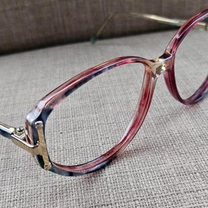 Könnte beinhalten: Eine Brille mit rosafarbenem und weißem Muster und goldenen Akzenten. Die Brille hat eine abgerundete, ovale Form und einen dünnen Rahmen.