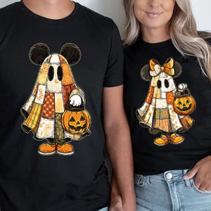 以下が含まれることがあります： ミッキーマウスの耳を持つ漫画の幽霊のキャラクターが描かれた黒いTシャツ。ジャック・オ・ランタンを持っています。幽霊は、オレンジと黄色のアクセントが付いたパッチワークのデザインで、ハロウィーンに最適です。