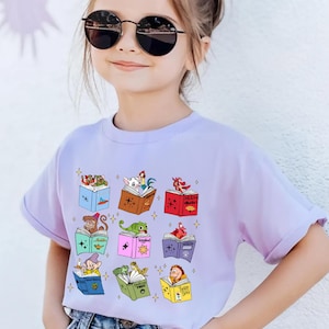 Puede incluir: Una camiseta de color lila claro con un gráfico colorido de animales y personajes de dibujos animados leyendo libros. El diseño incluye un mono, un dragón y otras figuras caprichosas. La camiseta la lleva una persona joven con gafas de sol.