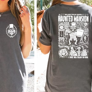 May include: A dark gray t-shirt with a white graphic print of the Haunted Mansion attraction at Disney World. The front of the shirt features a ghost with a top hat. The back of the shirt features a detailed illustration of the Haunted Mansion with text that reads "Welcome Foolish Mortals Haunted Mansion 999 Haunts Your Ghost Host Call on the Spirits in the Mansion A Ghost Will Follow You Home."