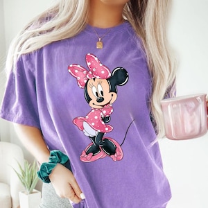 May include: Purple t-shirt featuring a graphic of Minnie Mouse wearing a pink polka dot dress and a red bow.