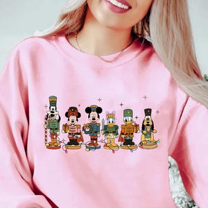 May include: A light pink sweatshirt featuring a line of cartoon nutcracker characters. The characters are dressed in festive outfits and stand on a string of lights. The design includes Mickey Mouse, Donald Duck, and Goofy. A Christmas tree is visible in the background.