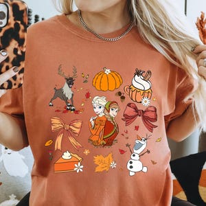 May include: Rust-colored t-shirt featuring a collection of fall-themed illustrations. The design includes a reindeer, pumpkins, pie, bows, autumn leaves, and characters from the movie Frozen. The shirt is a comfortable, casual style.
