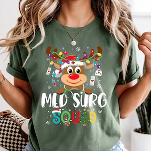 Puede incluir: Camiseta verde con un gráfico de reno con gorro de Papá Noel y equipo médico. El texto "MED SURG SQUAD" se muestra debajo del reno. La camiseta está hecha de un material suave y es adecuada para uso casual.