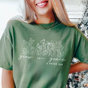 May include: A moss green t-shirt with the words "grow in grace" and the Bible verse 2 Peter 3:18 printed in white. The shirt features a floral and butterfly design. The shirt is made of a soft material.