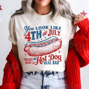 May include: A white t-shirt with a red, white, and blue graphic that says "You look like the 4th of July. Makes me want a Hot Dog real bad." The graphic features a hot dog with mustard and ketchup.