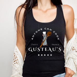 May include: Black tank top with a white graphic of a rat chef holding a wooden spoon. The graphic says "Anyone Can Cook" with "Paris" and "France" on either side of the rat. The graphic also says "Gusteau's" with five stars below the text.