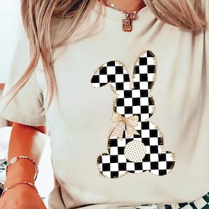 Op de afbeelding: Een wit T-shirt met een zwart-wit geruit konijnontwerp. Het konijn heeft een gouden strik en een gouden stippenpatroon op zijn buik.
