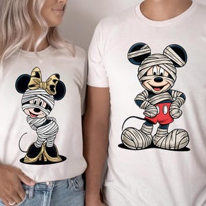 May include: White t-shirts featuring cartoon characters dressed as mummies. One shirt has a mummy Minnie Mouse with a gold bow, and the other has a mummy Mickey Mouse in red shorts. The word "WHITE" is at the bottom.