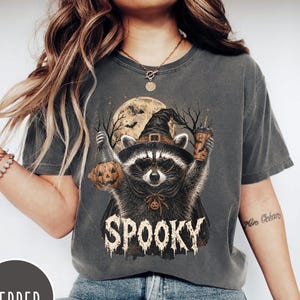 Pu&ograve; includere: Una t-shirt a maniche corte grigio antracite con la grafica di un procione che indossa un cappello da strega e occhiali da sole. Il procione tiene in mano una zucca e un bicchiere di caff&egrave; sotto una luna piena. La scritta SPOOKY &egrave; in bianco.