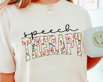 May include: A white t-shirt with the text "speech THERAPY" in black lettering. The text is surrounded by a floral pattern in pink, green, and yellow.