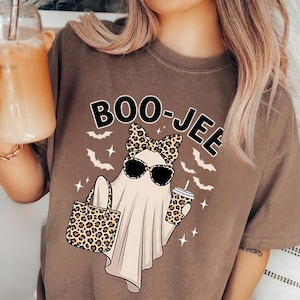 May include: Espresso-coloured t-shirt with a cartoon ghost wearing sunglasses and a leopard print bow, holding a leopard print tote bag and a coffee cup. The text "BOO-JEE" is above the ghost.