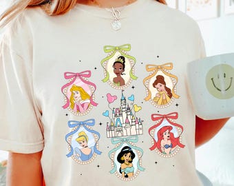 Peut inclure: Un t-shirt blanc avec un motif de princesses Disney dans des cadres avec des nœuds. Les princesses incluent Cendrillon, Ariel, Jasmine, Tiana, Belle et Blanche-Neige. Le motif comprend également une représentation du château de Cendrillon.