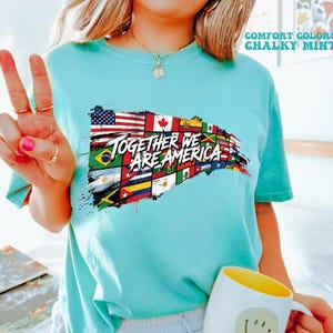 May include: A light blue t-shirt with the text "TOGETHER WE ARE AMERICA" over a design of various national flags. The shirt also has the words "COMFORT COLORS" and "CHALKY MINT" printed on it. A white mug with a yellow interior and a smiley face is also visible.