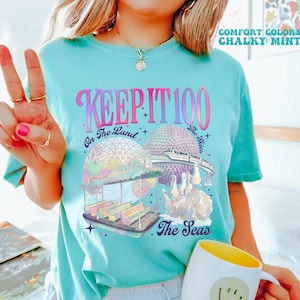 May include: A light blue t-shirt with the text "KEEP IT 100" in a pink and purple font. The shirt features a colorful graphic of a theme park with the text "On The Land The Skies The Seas". A white mug with a yellow interior and a smiley face is also visible.