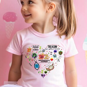 Puede incluir: Una niña pequeña con una camiseta rosa claro con un gráfico en forma de corazón. El gráfico incluye las palabras "Proud Zombies" y "Treason she's guilty of treason" junto con otras ilustraciones coloridas. El fondo es una pared rosa con diseños de conos de helado.