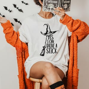 Pu&ograve; includere: T-shirt bianca con cappello da strega e grafica scopa, il testo recita "Yes I Can Drive a Stick". La persona indossa un cardigan arancione e calzini a tema zucca. Viene tenuto in mano un libro intitolato "Spells & Potions".