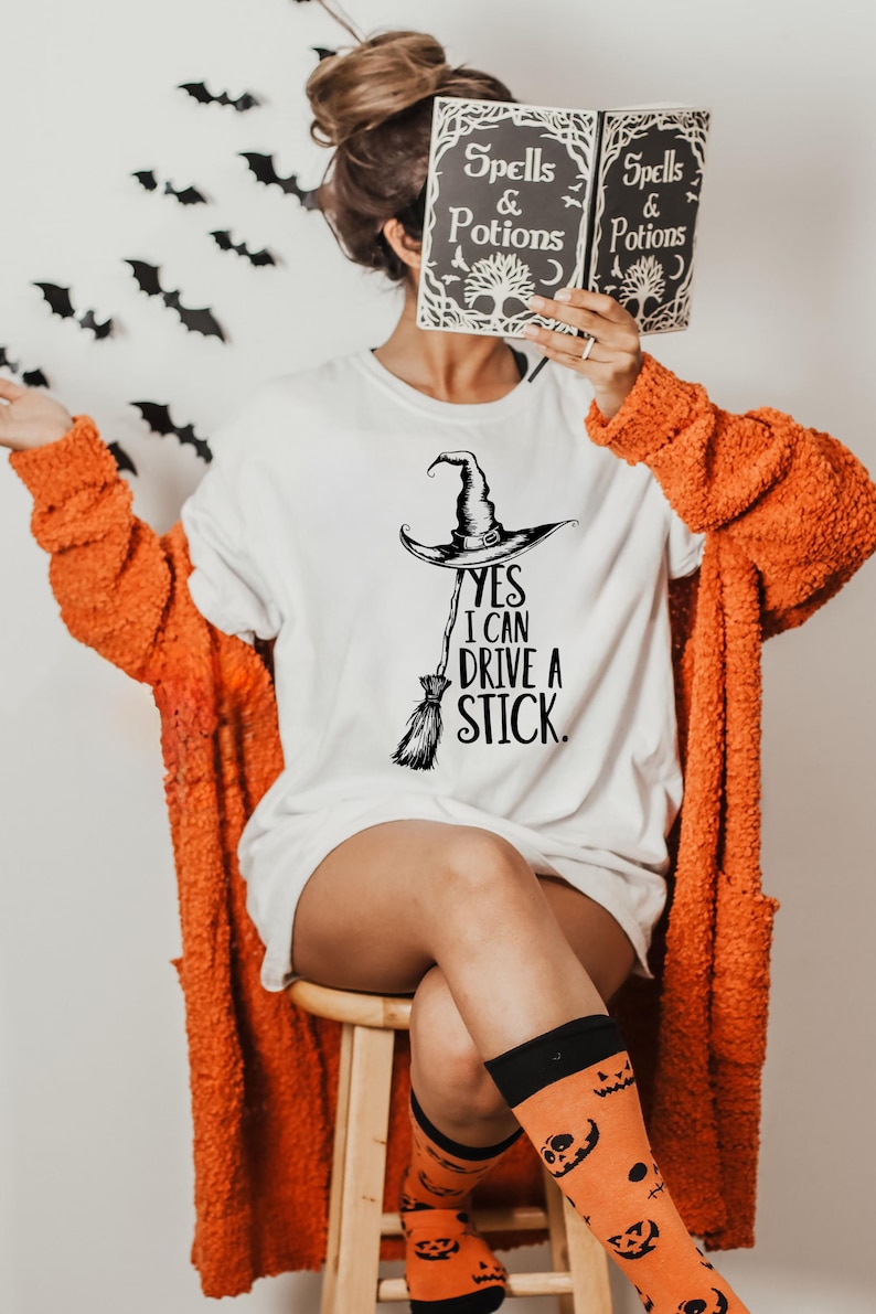 Pu&ograve; includere: T-shirt bianca con cappello da strega e grafica scopa, il testo recita "Yes I Can Drive a Stick". La persona indossa un cardigan arancione e calzini a tema zucca. Viene tenuto in mano un libro intitolato "Spells & Potions".