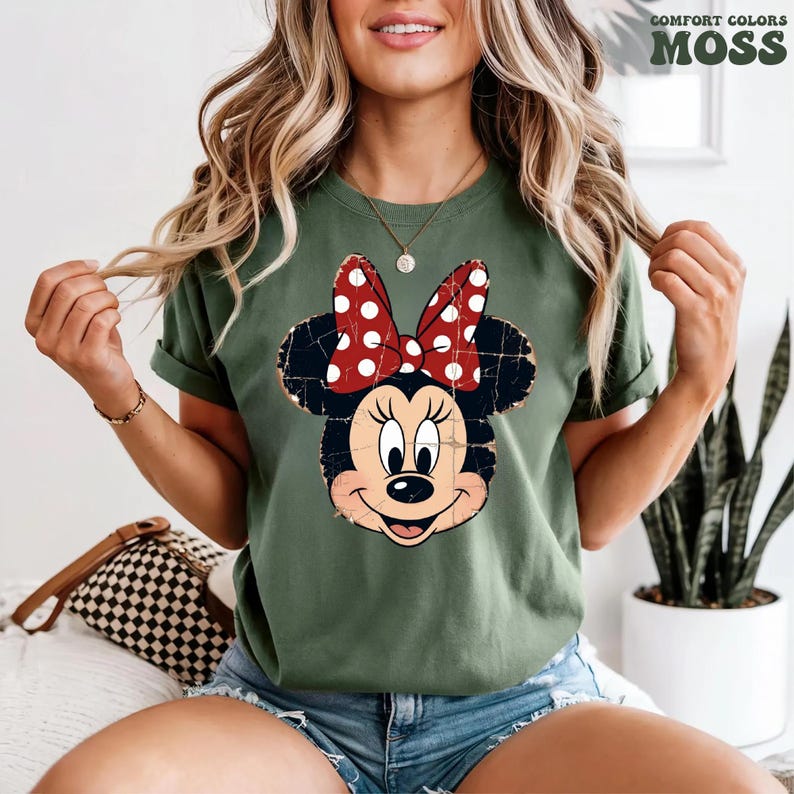 May include: A moss green short-sleeved t-shirt featuring a distressed graphic of Minnie Mouse with a red and white polka dot bow. The shirt has a crew neckline and a relaxed fit. The text COMFORT COLORS MOSS is printed in the top right corner.