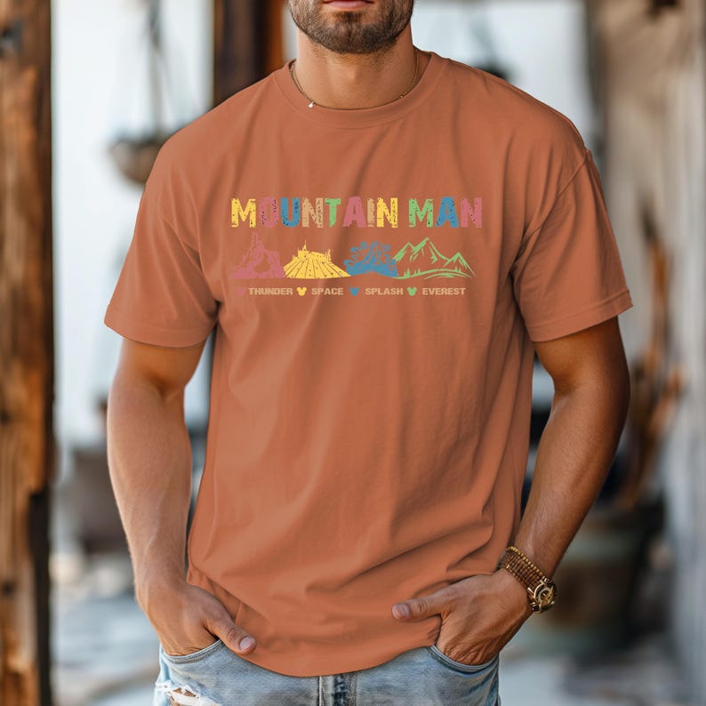 May include: A brown t-shirt with the text "Mountain Man" printed in colorful letters. The text is accompanied by a graphic of mountains and roller coasters. The text below the graphic reads "Thunder, Space, Splash, Everest".