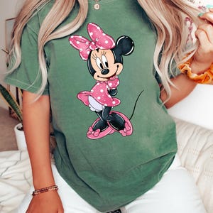 May include: A green t-shirt featuring a cartoon image of Minnie Mouse wearing a pink polka dot dress and a pink bow.