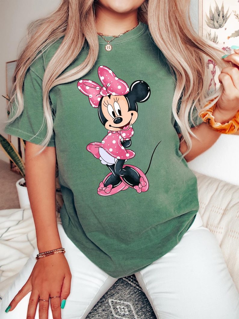 May include: A green t-shirt featuring a cartoon image of Minnie Mouse wearing a pink polka dot dress and a pink bow.