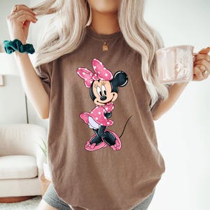 May include: A brown t-shirt featuring a graphic of Minnie Mouse wearing a pink polka dot dress and a pink bow.