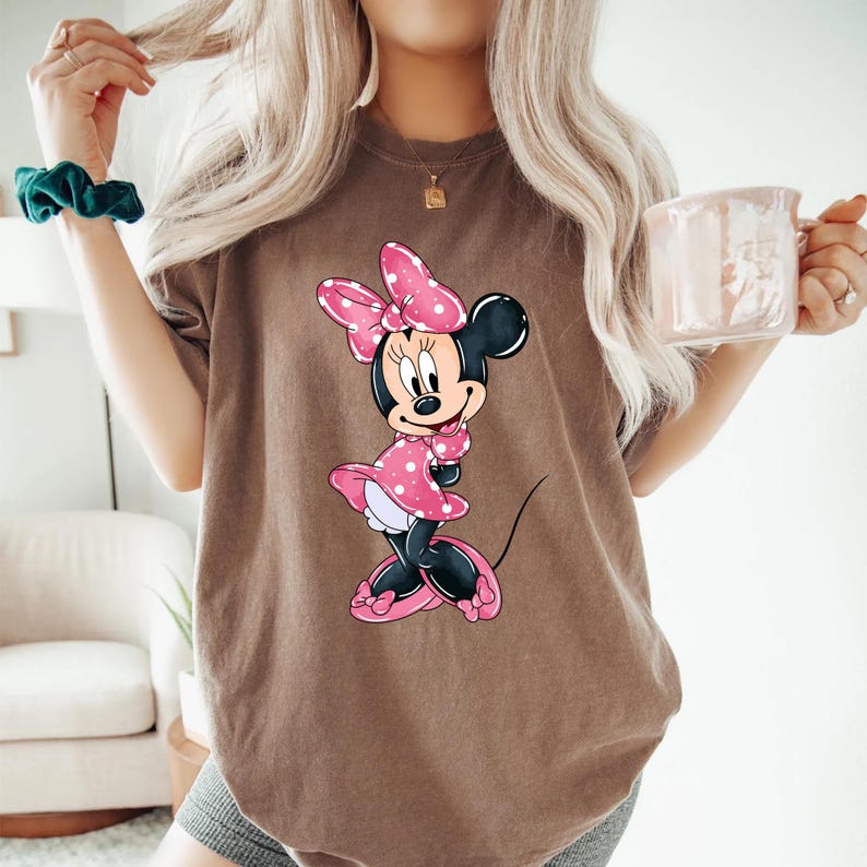 May include: A brown t-shirt featuring a graphic of Minnie Mouse wearing a pink polka dot dress and a pink bow.