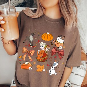 May include: A brown t-shirt featuring a colorful graphic with cartoon images of a reindeer, pumpkin, sisters, a cup of coffee, bows, pie, and Olaf the snowman. The design has a fall or autumn theme.
