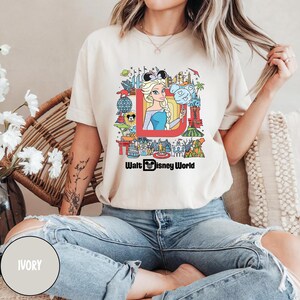 Może przedstawiać: Biały t-shirt z kolorową grafiką przedstawiającą księżniczkę Disneya i r&oacute;żne atrakcje Disneya. Pod grafiką znajduje się napis "Walt Disney World". Koszulkę nosi osoba z tatuażem na ramieniu. Kolor koszulki to kość słoniowa.