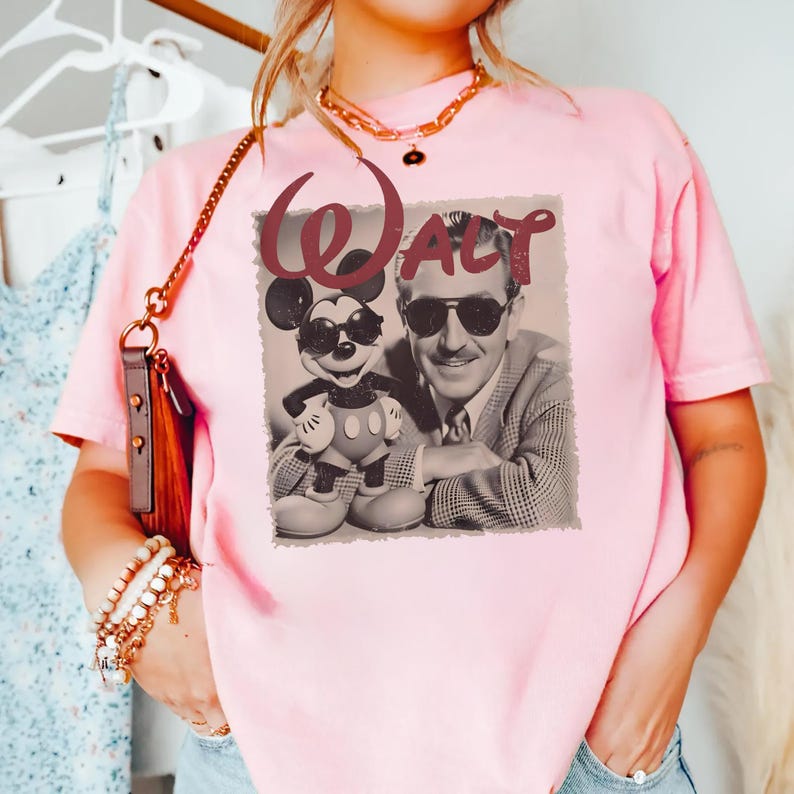Op de afbeelding: Een lichtroze T-shirt met korte mouwen en een grafische print van Walt Disney en Mickey Mouse die beide een zonnebril dragen. De tekst "Walt" is in een donkerrood cursief lettertype boven de figuren op een rechthoekige achtergrond gedrukt.