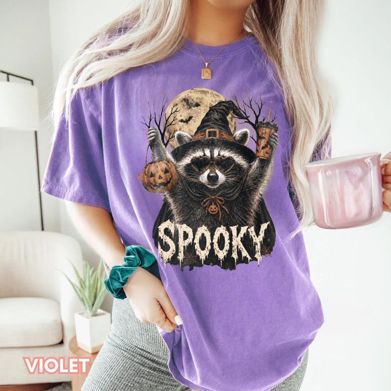 Pu&ograve; includere: Una t-shirt viola a maniche corte con la grafica di un procione che indossa un cappello da strega e occhiali da sole a forma di cuore. Il procione tiene in mano una zucca e un bicchiere. Sullo sfondo luna piena e alberi con il testo SPOOKY.